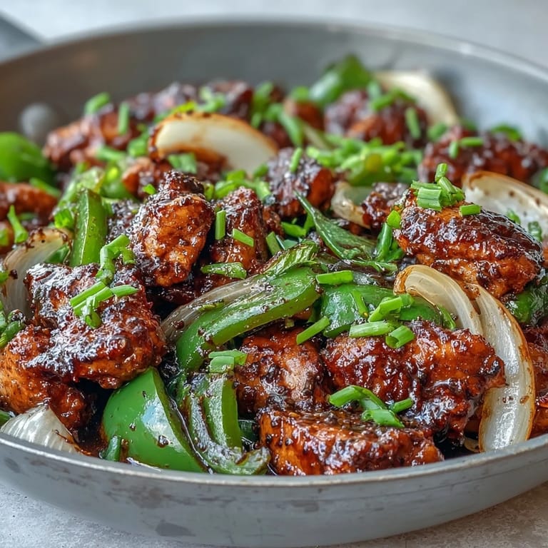 A close-up of Black Pepper Chicken reveals juicy chicken pieces and fresh aromatics in a rich, glossy sauce.